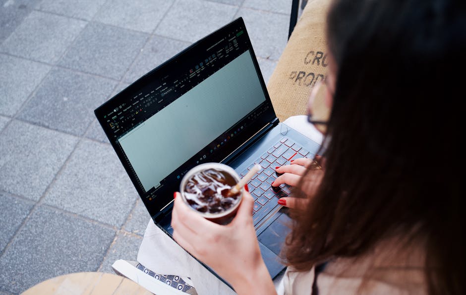 A woman works on a laptop outdoors, holding an iced drink with a straw in a disposable cup.