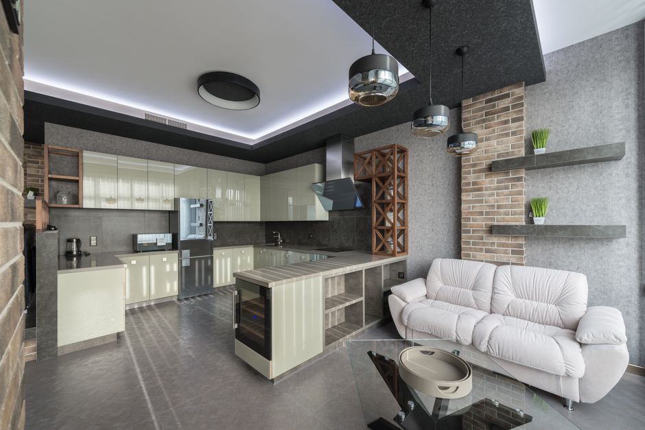 White leather sofa placed near kitchen cupboards with modern appliances in light spacious kitchen zone with decorative elements in apartment